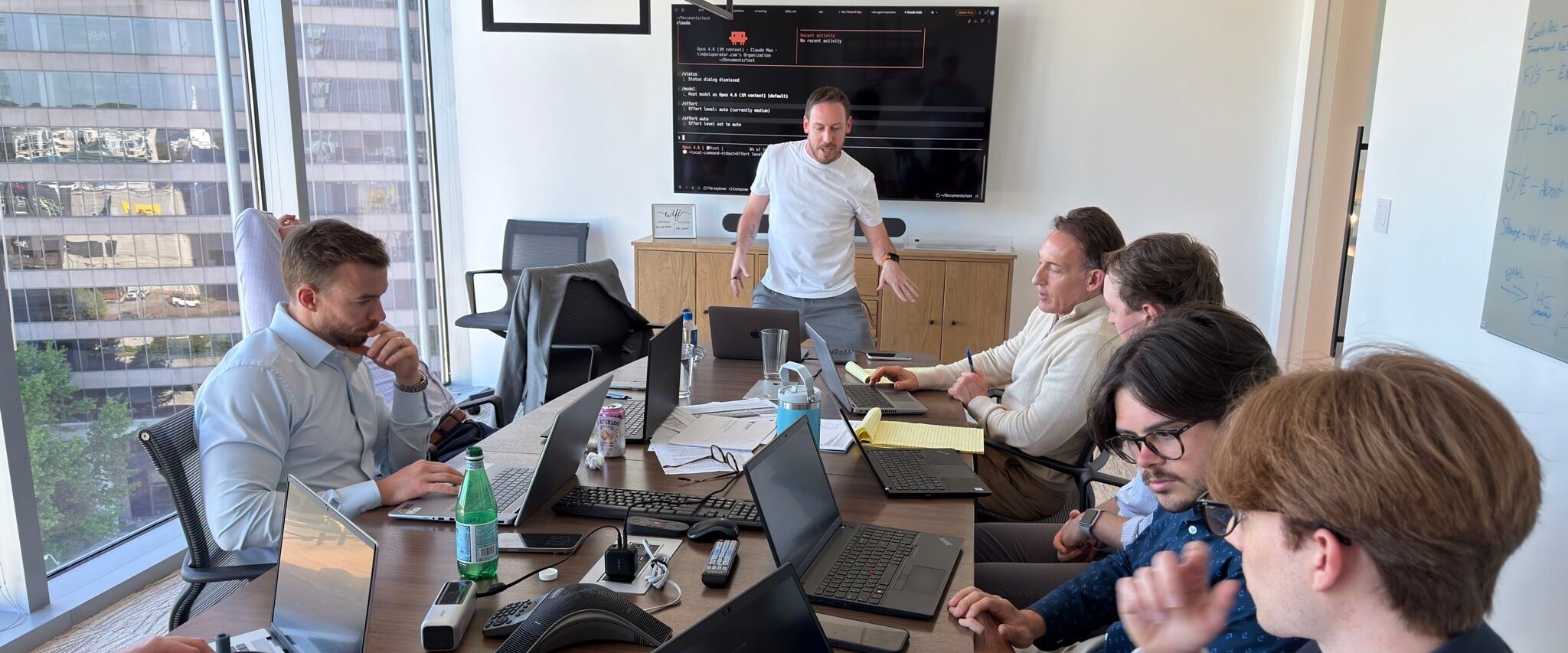 Tim Cakir standing at the head of a client boardroom table, leading a session for six people working on laptops, with Claude Code open on the wall screen behind him.