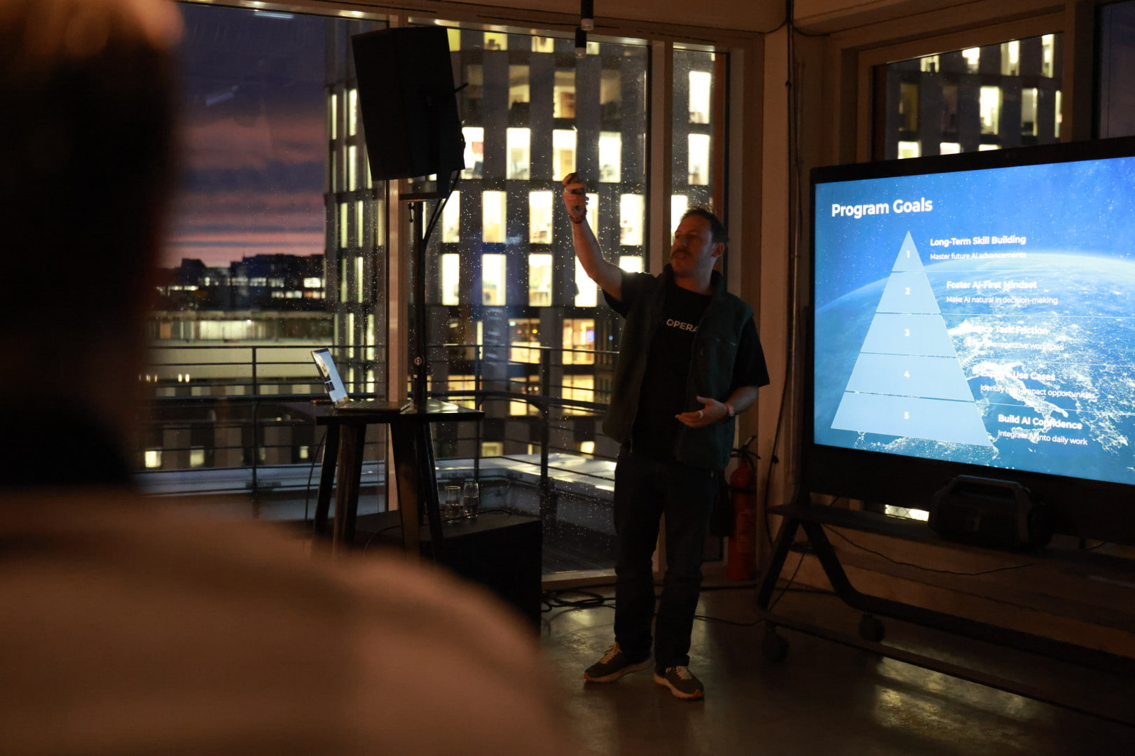 Tim Cakir presenting to a small group at a client kick-off in Munich, one hand raised towards a screen headed Program Goals, with city lights and rain on the windows behind him.