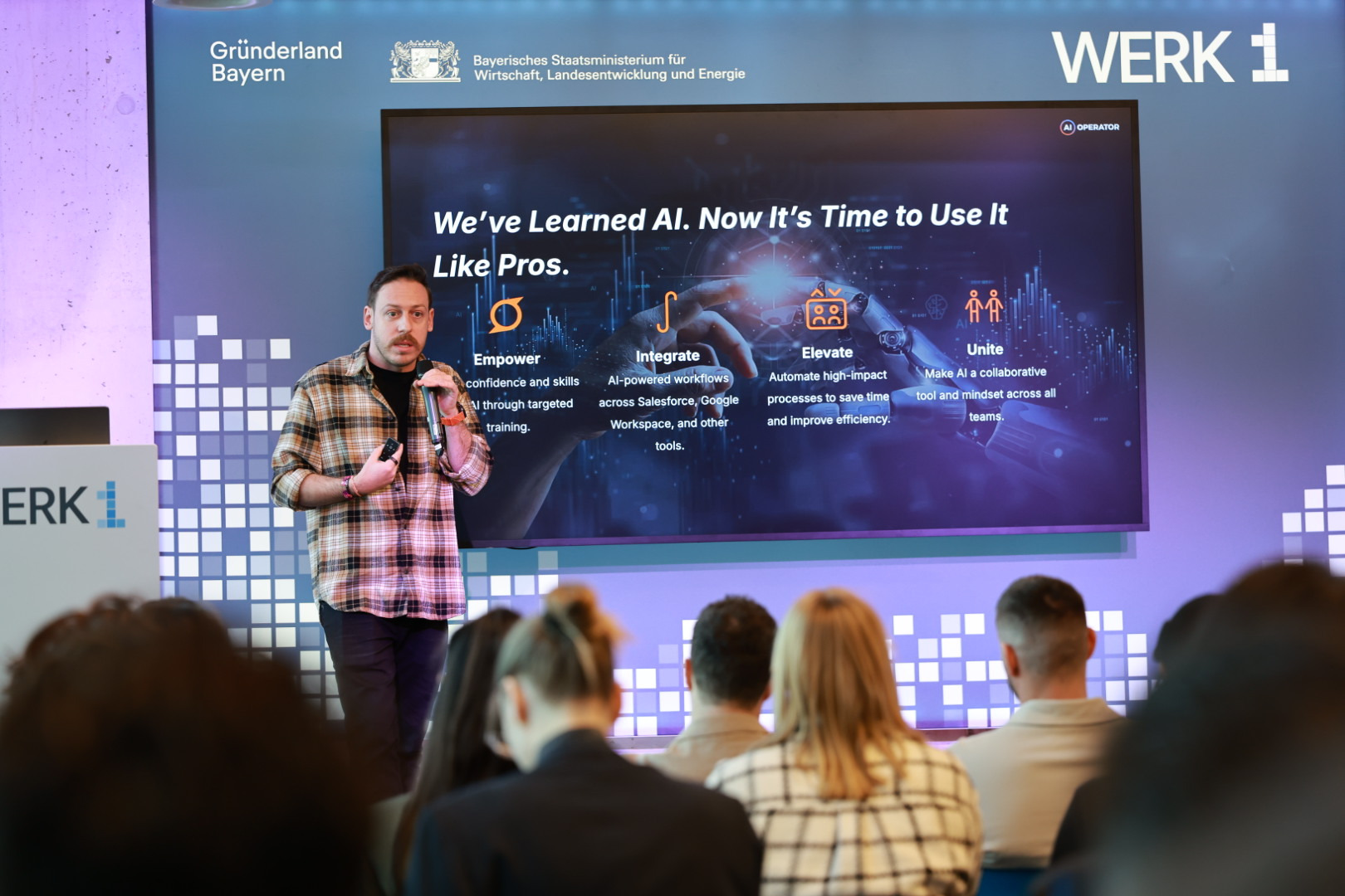 Tim Cakir on stage holding a microphone at a client kick-off, a large screen behind him reading We've Learned AI. Now It's Time to Use It Like Pros, with the audience seated in front of him.